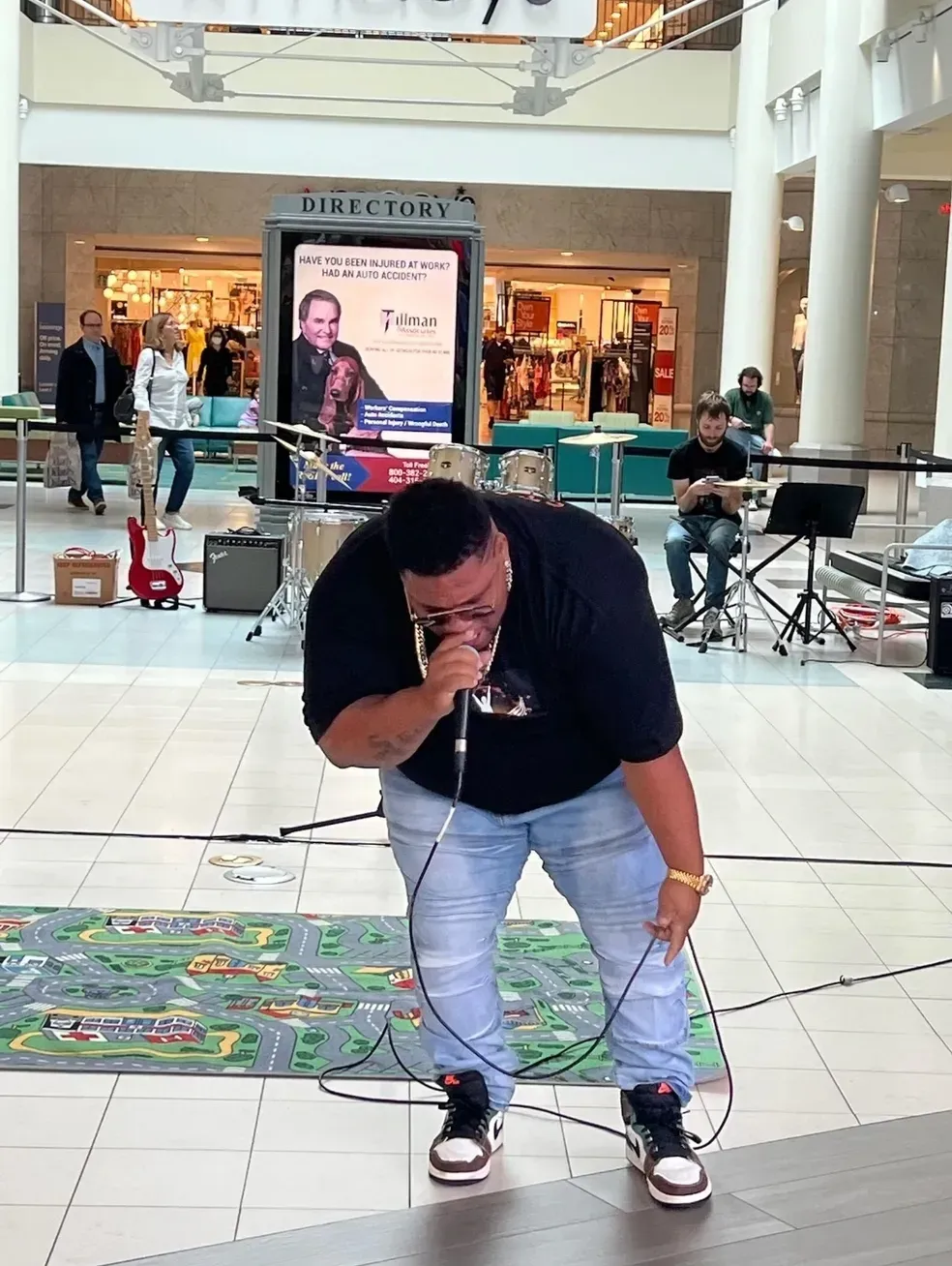 Man singing into a microphone in a mall atrium, bent forward on a tiled floor with a colorful mat.