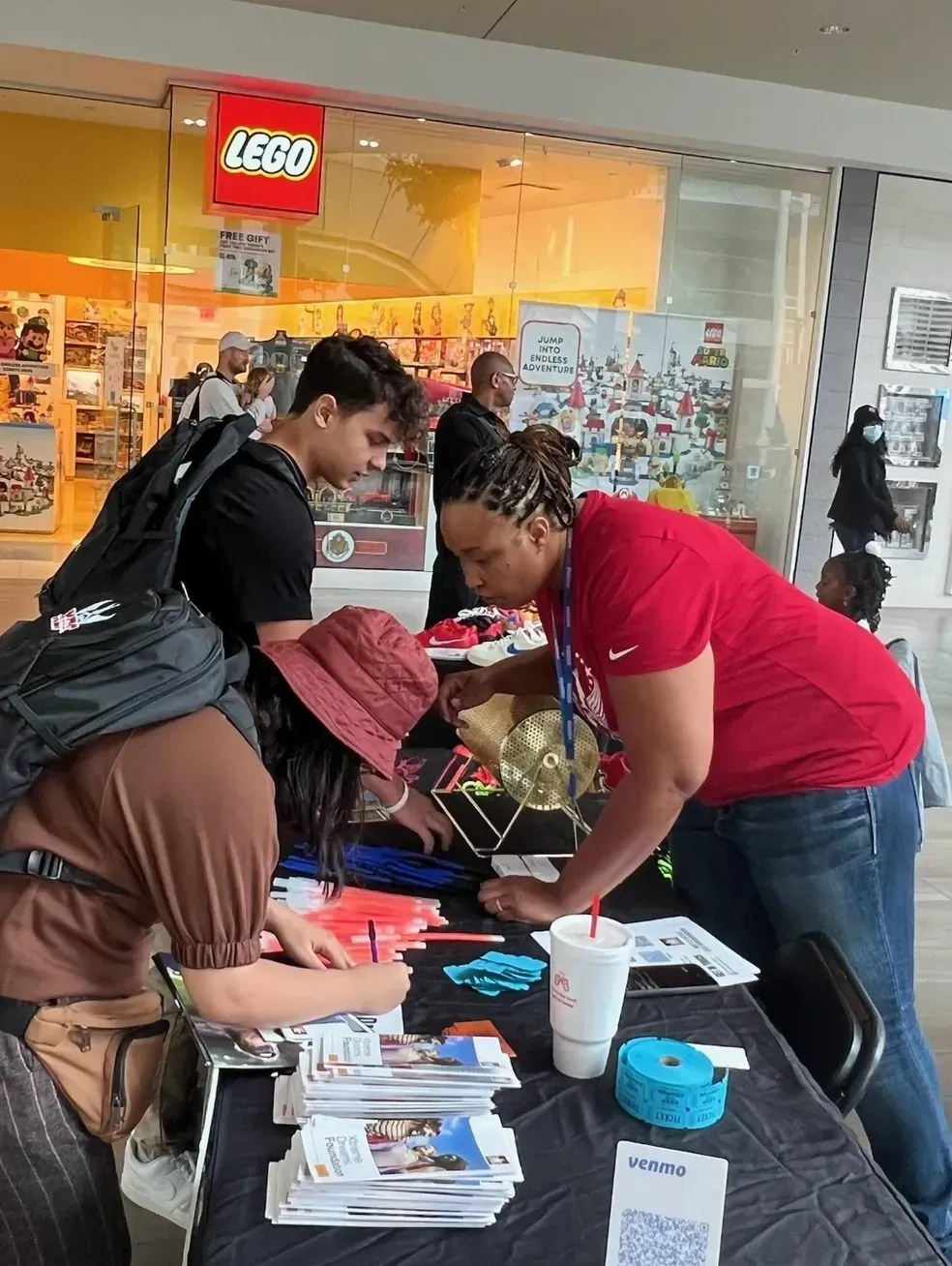 Volunteers assisting people at a table inside a mall, with forms, cups, and supplies spread out.