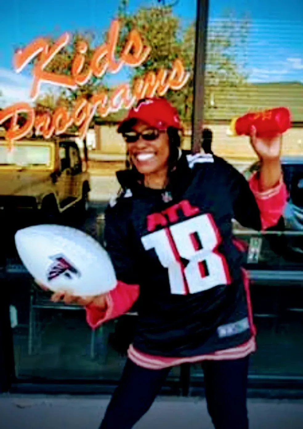 Woman in black football jersey and red cap posing outside Kids Program storefront, holding a football and foam finger