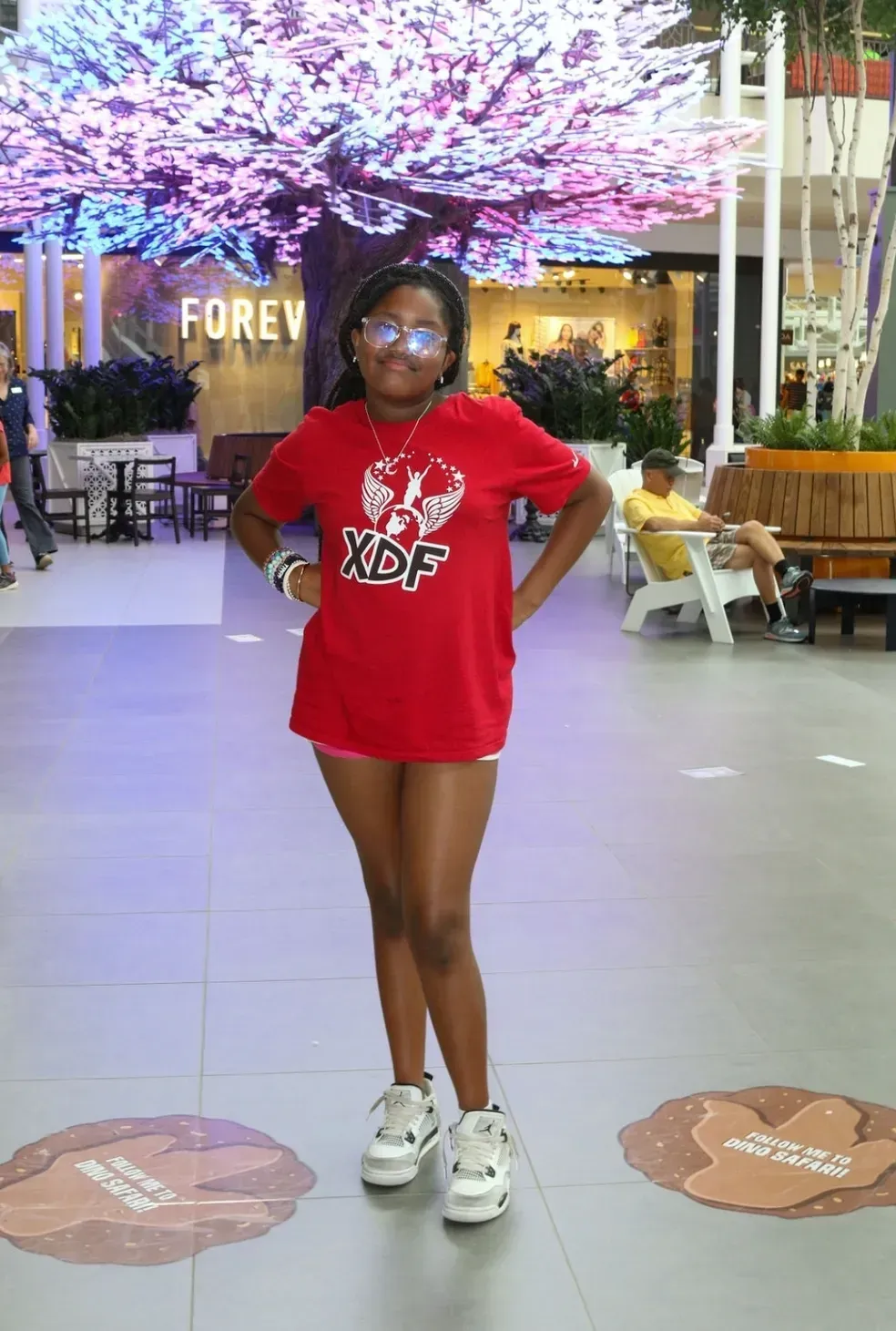 Person in red shirt and white sneakers standing indoors with hands on hips, under pink-lit trees.