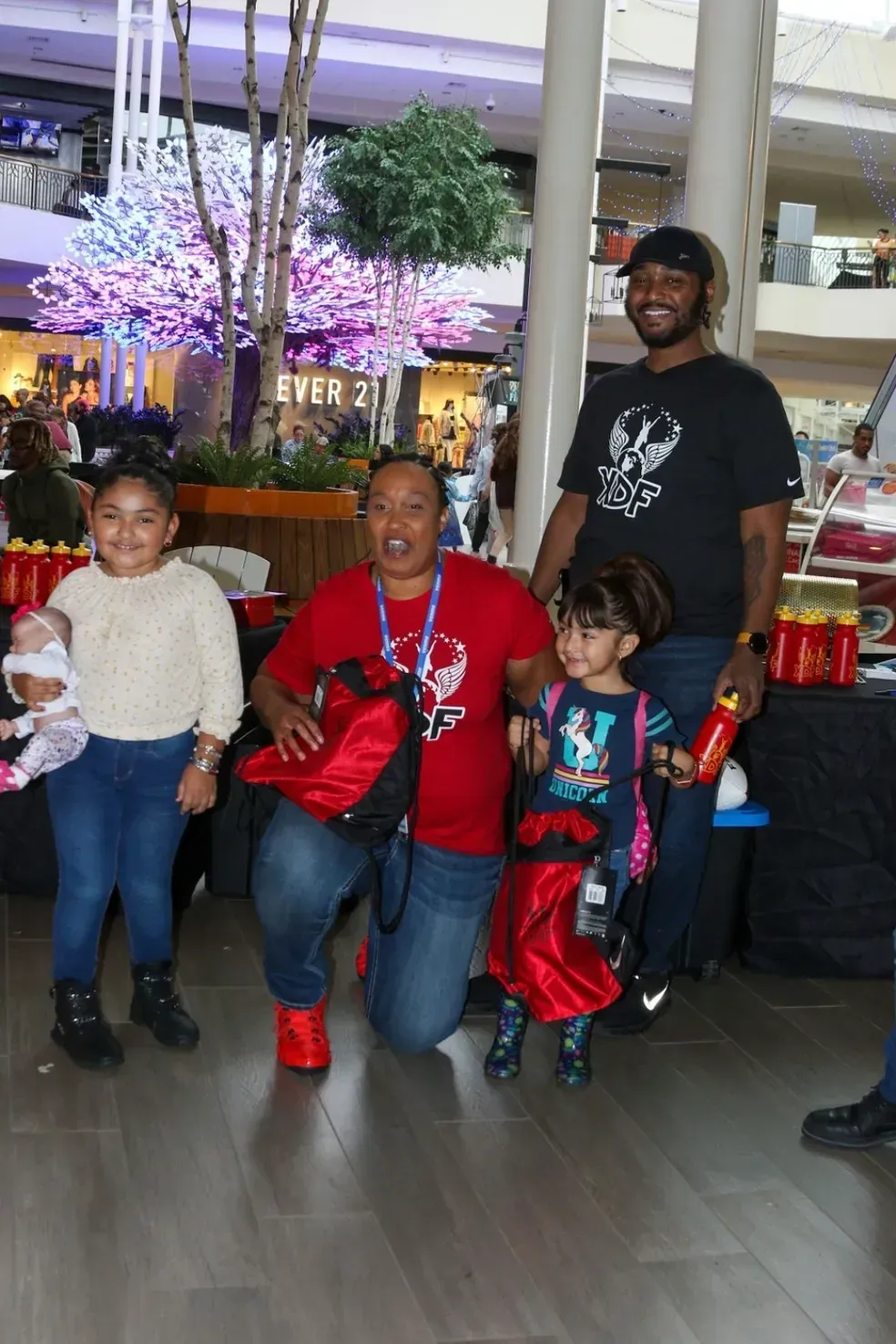 Four people pose indoors at a festive event, with two children in colorful costumes and purple-lit decor behind them