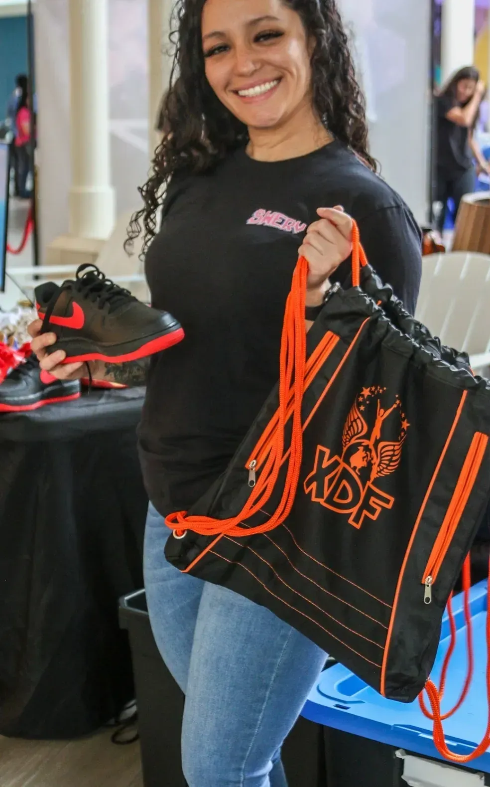Smiling woman holding a black-and-orange shoe and matching tote bag at a product display booth
