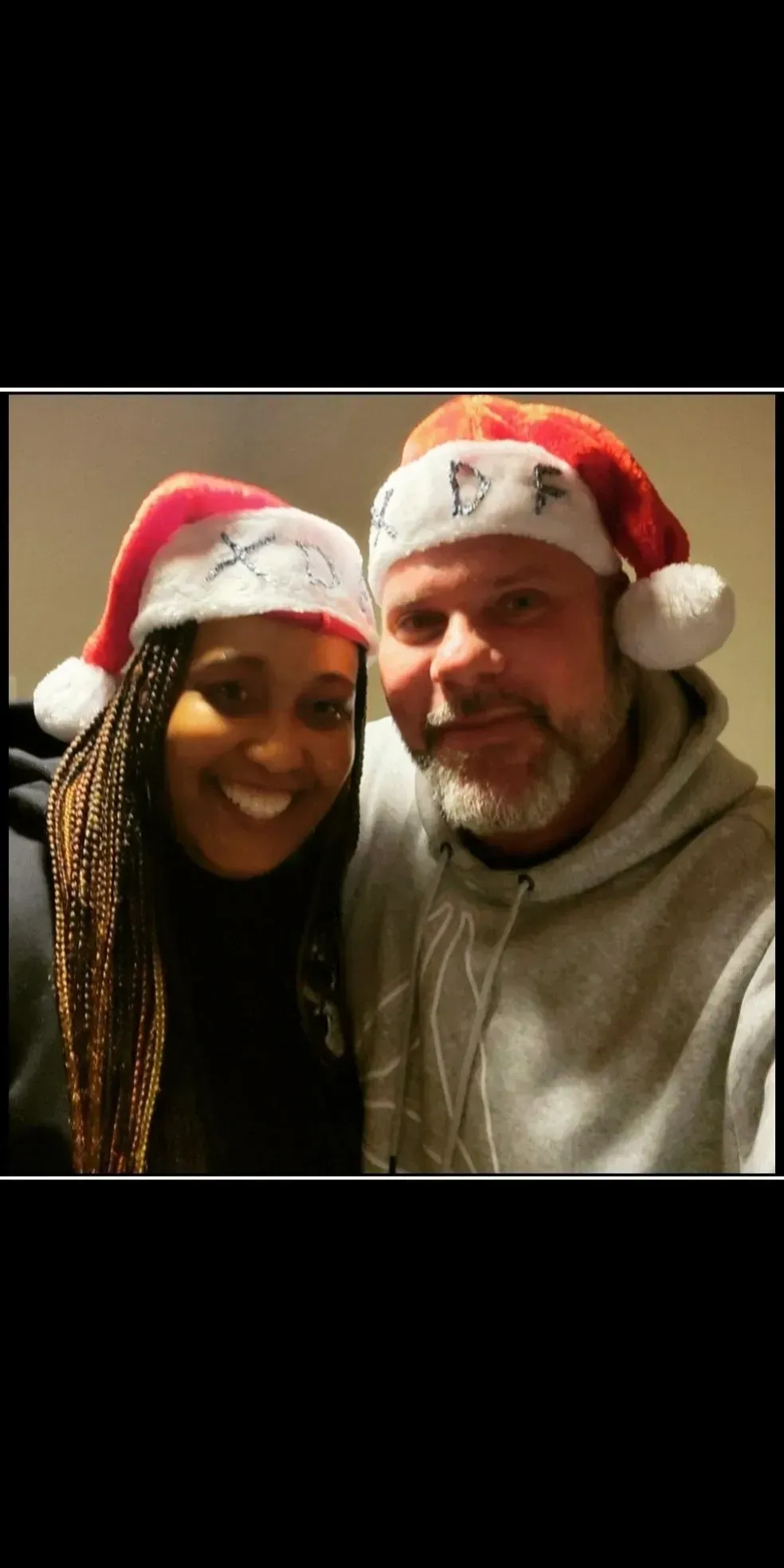 Two smiling people wearing red Santa hats pose indoors against a dark background.