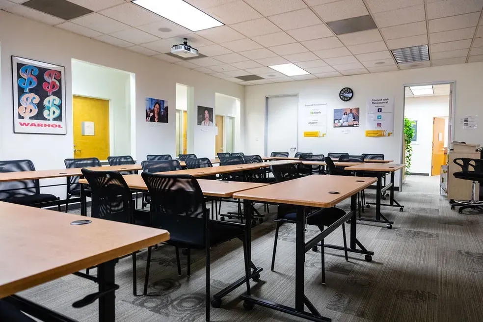 Empty classroom with desks, chairs, white walls, colorful posters, and daylight through windows