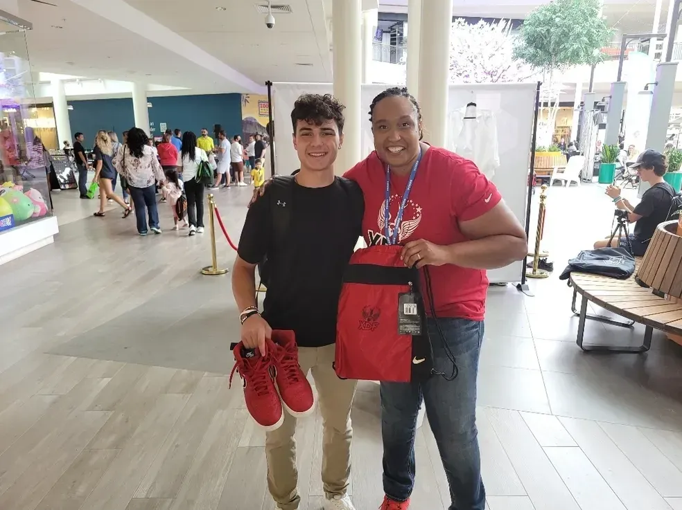 Two people smiling indoors, holding red shoes and a red shopping bag in a mall.