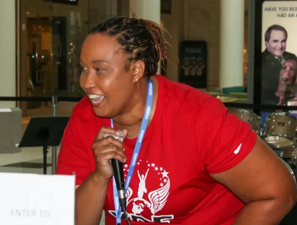 Woman in red shirt speaking into a microphone at an indoor event, smiling beside a sign.