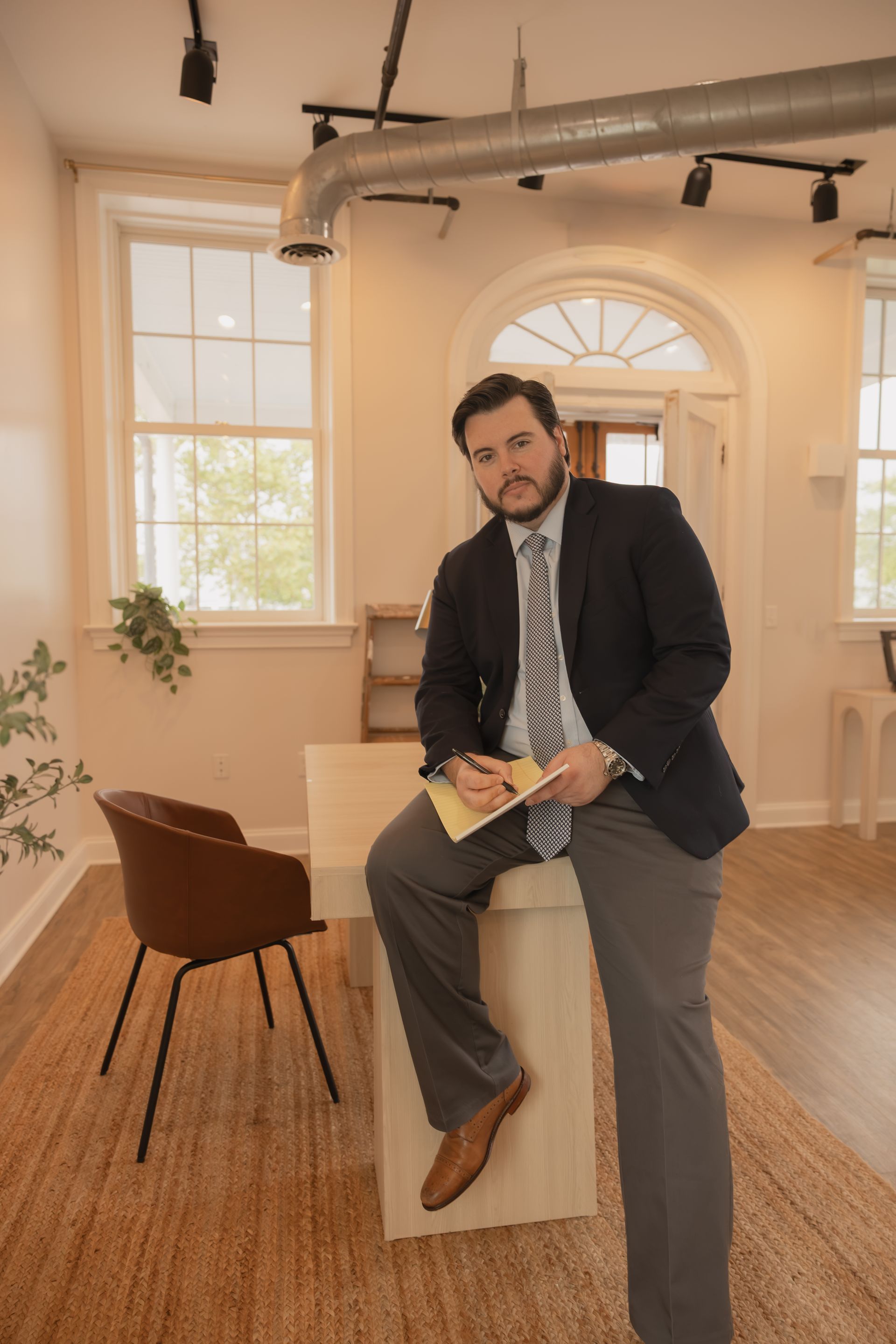 Man in a suit sitting on a box in a bright office, holding a notebook.