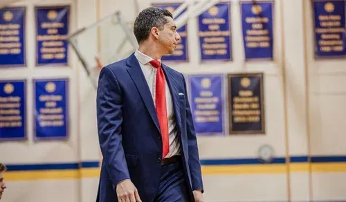 Man in navy suit and red tie looking left in a gym with blue championship banners
