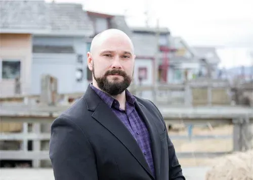 Bald man in a dark suit standing outdoors near a waterfront, with buildings in the background.