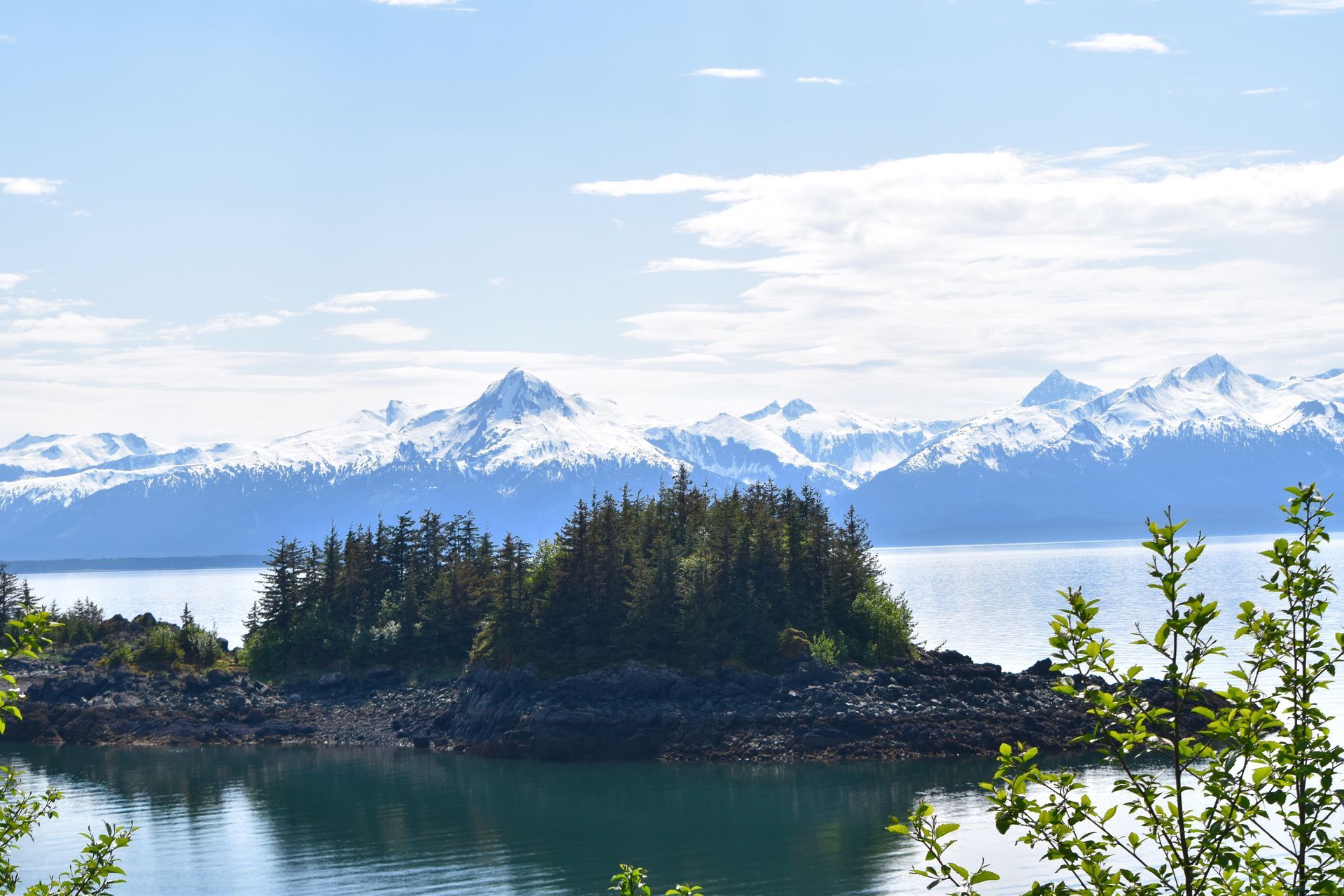 Snowy mountains above a forested island in calm blue water under a bright sky