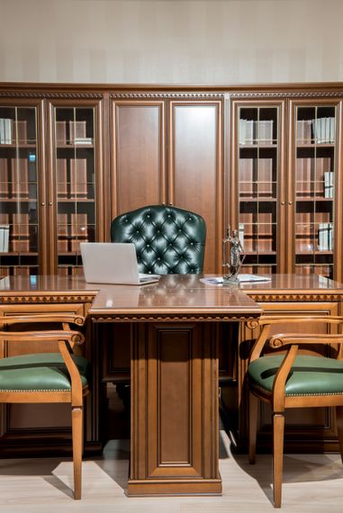 Wood-paneled library office with desk, laptop, green chairs, and empty green leather chair behind it