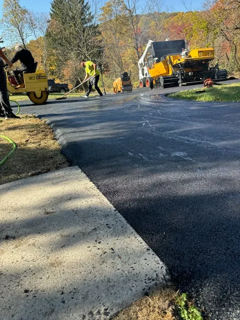 Road crew paving a street beside a sidewalk, with orange construction machinery and workers in autumn trees.