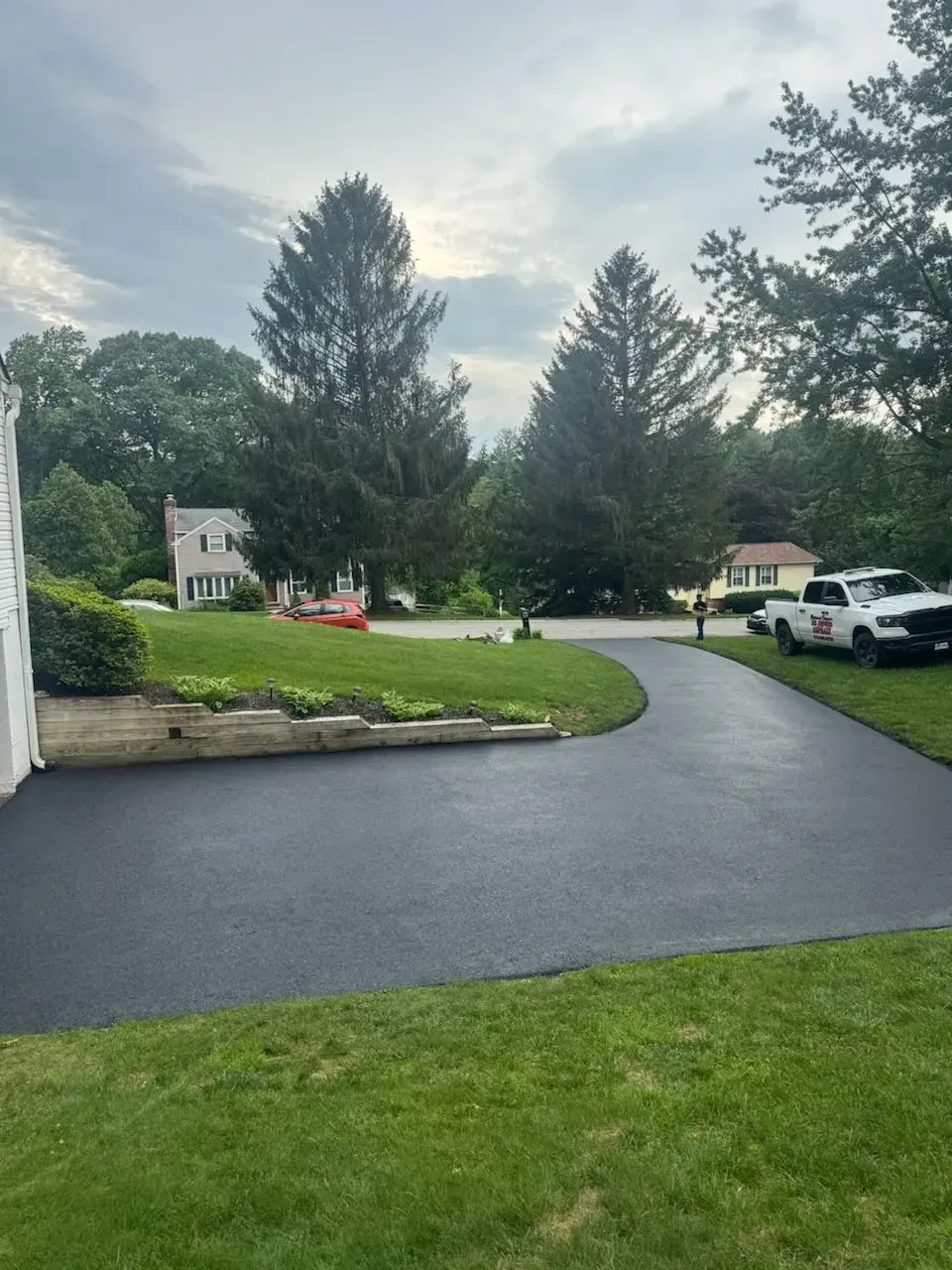 Curved black driveway beside a green lawn and trees in a suburban neighborhood after rain