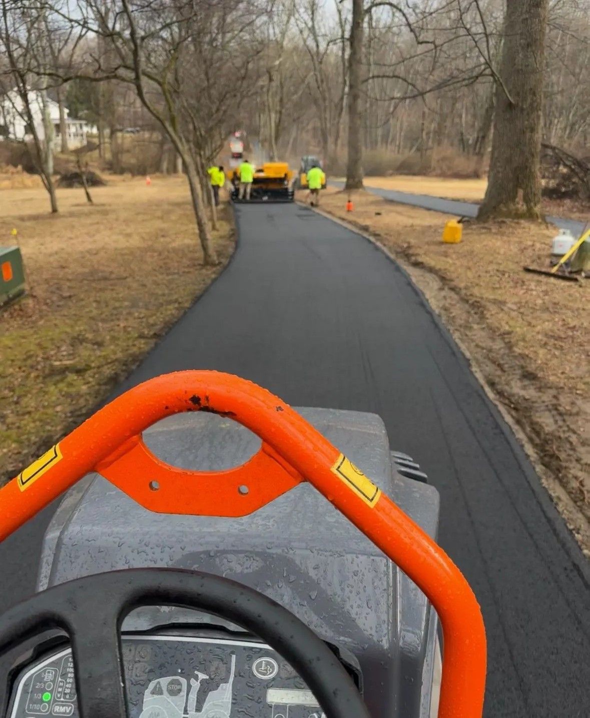 Worker on an orange machine paving a black path in a wooded park area