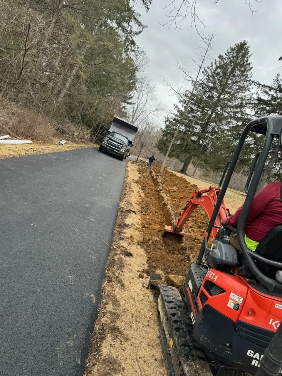 Small excavator digging a trench beside a newly paved road in a wooded area, with a truck ahead.
