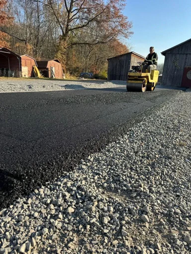Yellow road roller paving a gravel road beside barns and trees on a sunny day