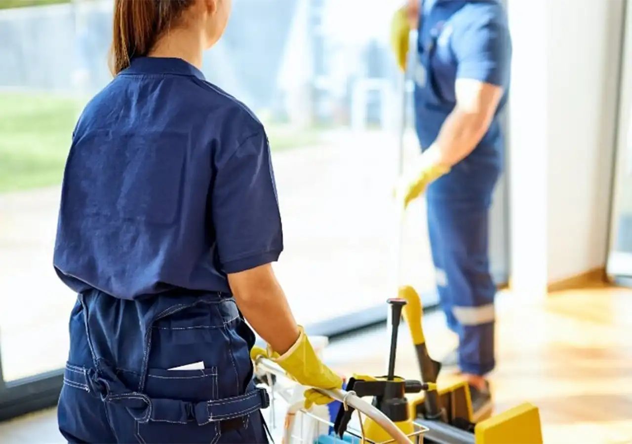 Two cleaning workers in blue uniforms cleaning a window, one with a cart, inside a building.
