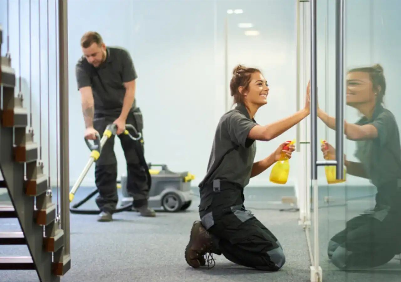 Two cleaning workers in an office; one vacuums, the other cleans a glass door, smiling.