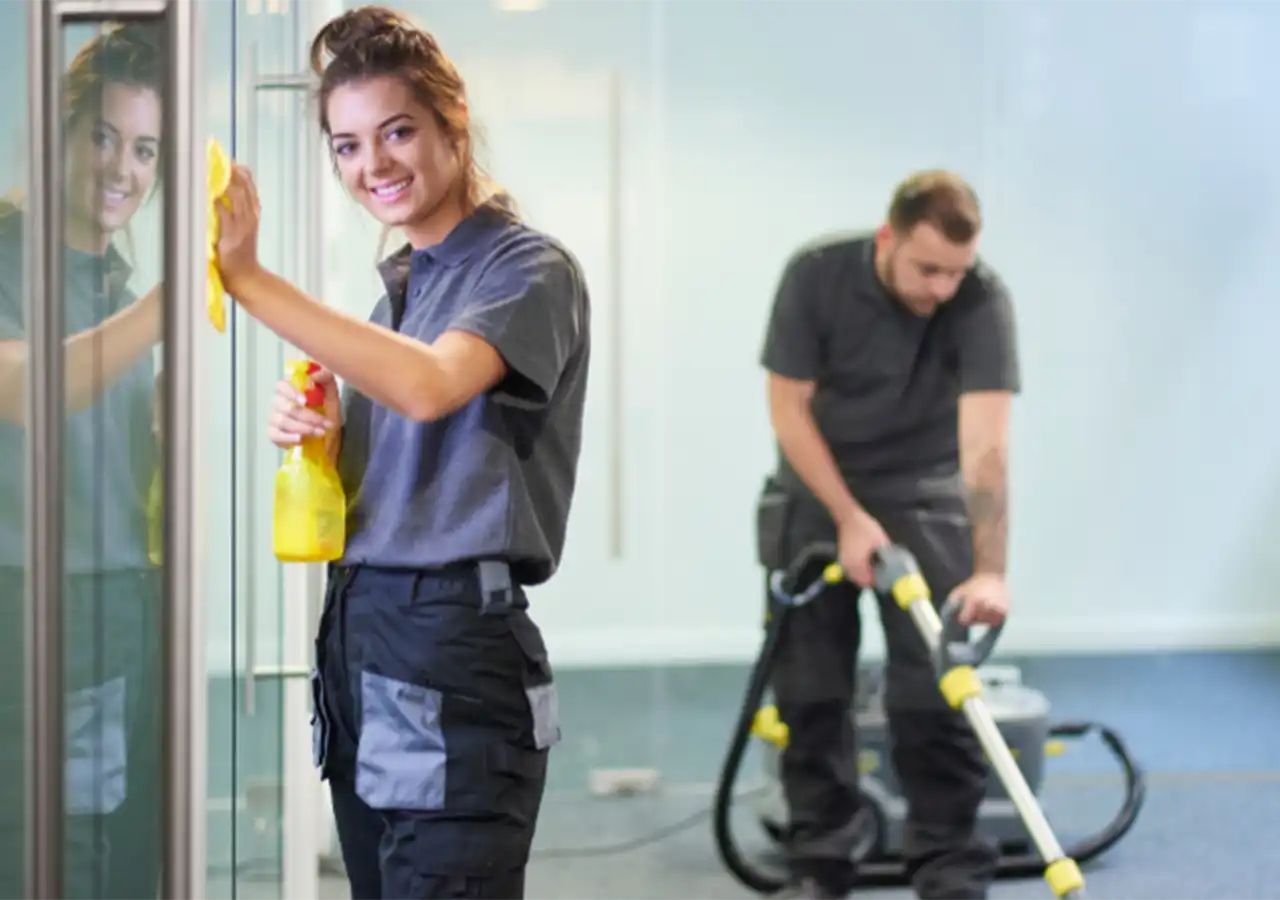 Woman cleaning glass door, smiling. Man vacuums office floor in background.
