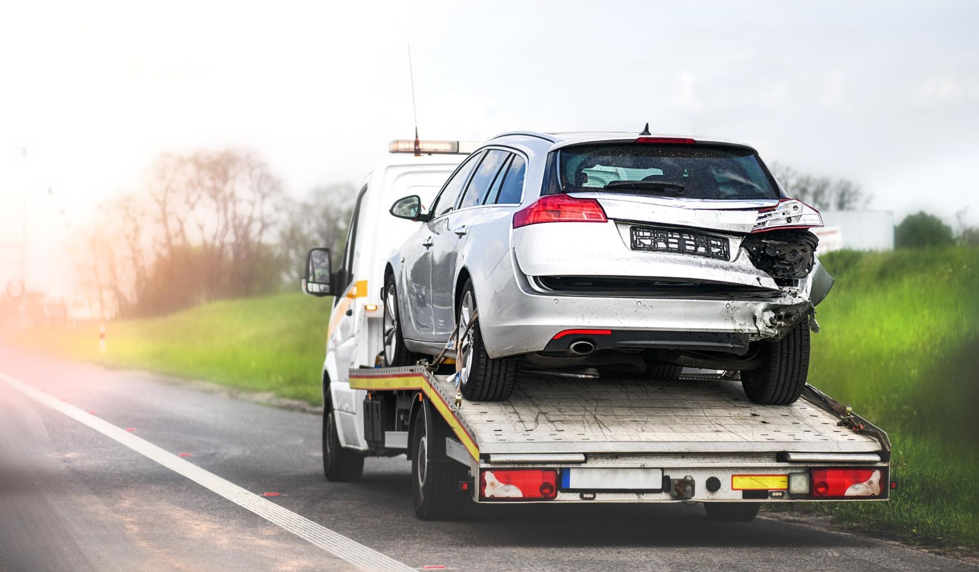 Silver car with rear-end damage on a tow truck, roadside setting.