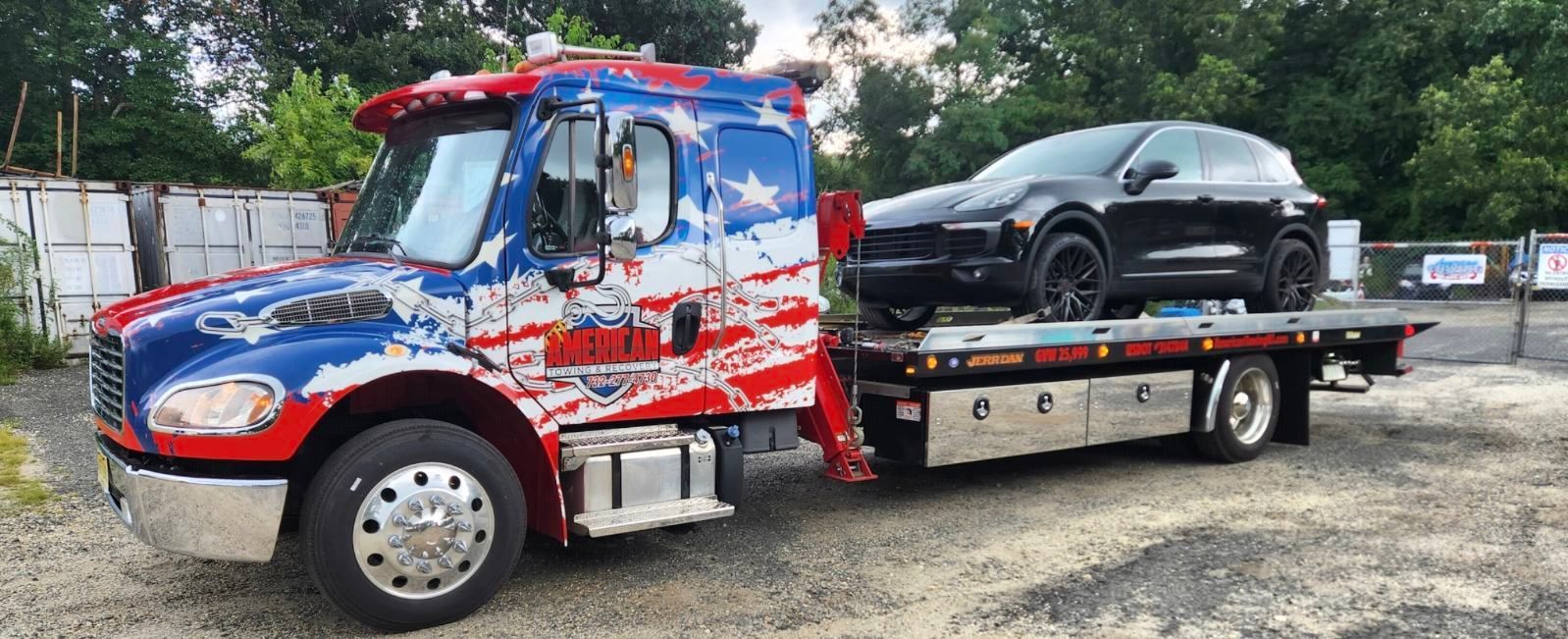 Tow truck with American flag paint job carrying a black car on its flatbed. Outdoors.
