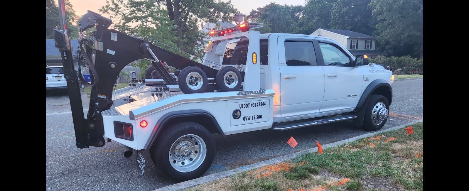 White tow truck parked on a roadside with its boom extended, with trees and a house in the background.