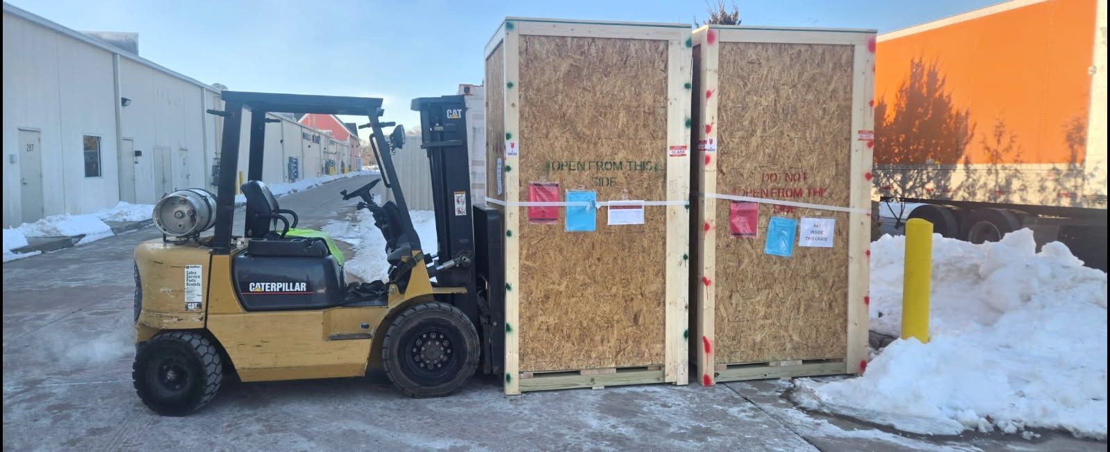 A yellow forklift carrying two large wooden crates outside a building with snow on the ground.