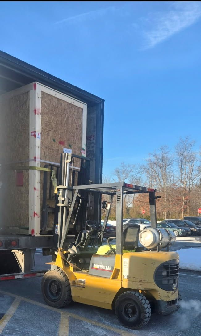 A yellow forklift loading a large wooden crate into a truck on a sunny day.