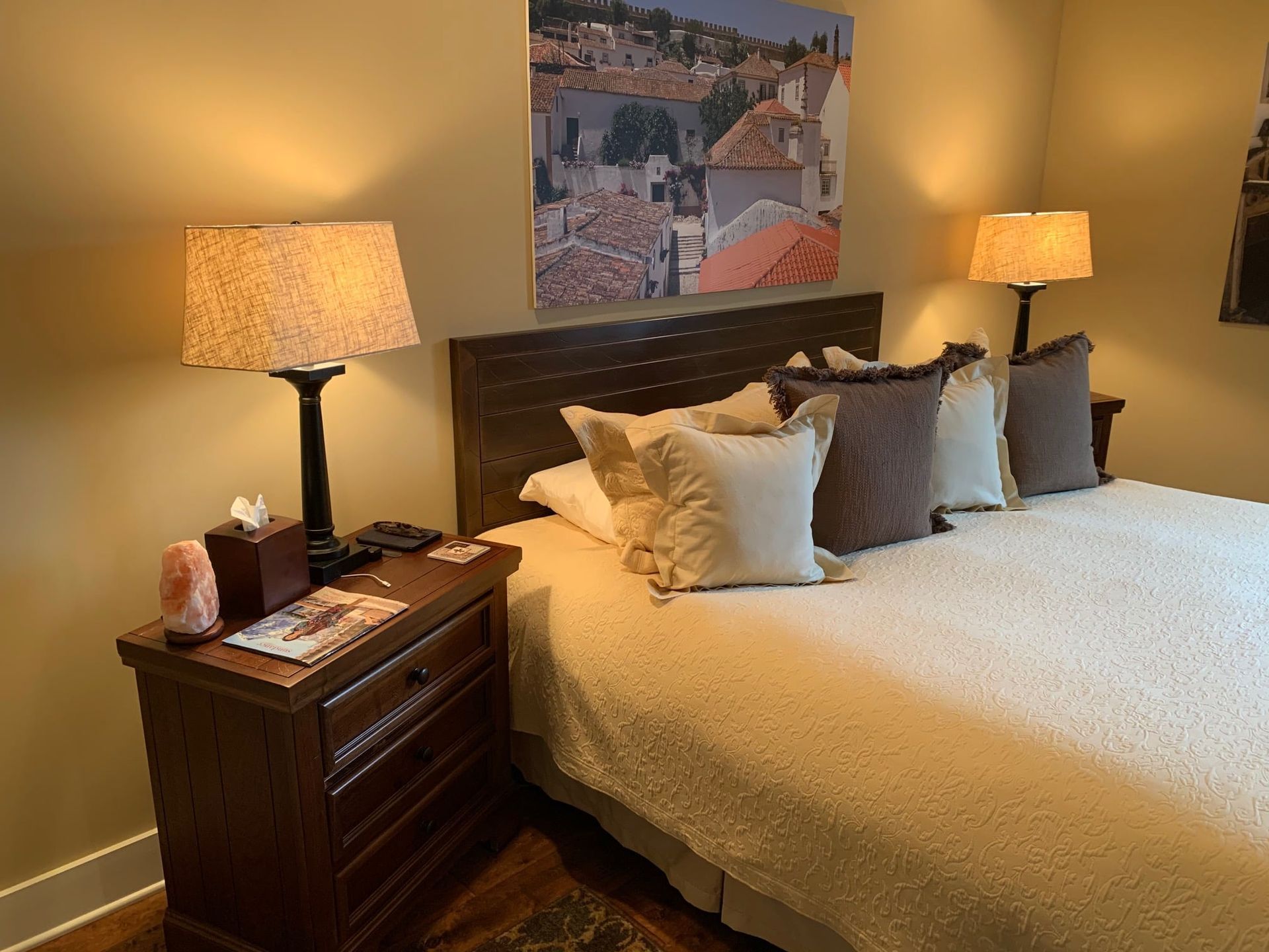 A king-sized bed with cream bedding, a dark wood headboard and nightstand, topped with a lamp and framed wall art.