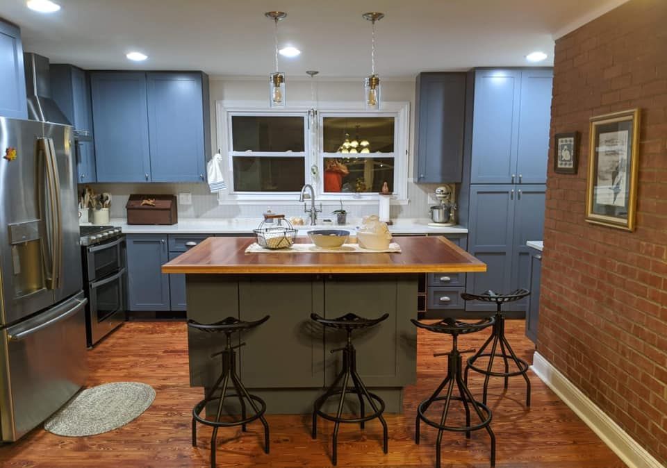 Modern kitchen with blue cabinets, a central wooden island with three metal bar stools, and a brick feature wall.