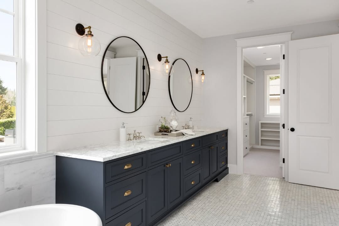 A modern bathroom features a long navy blue vanity with white counters, round mirrors, and wall sconces against white walls.