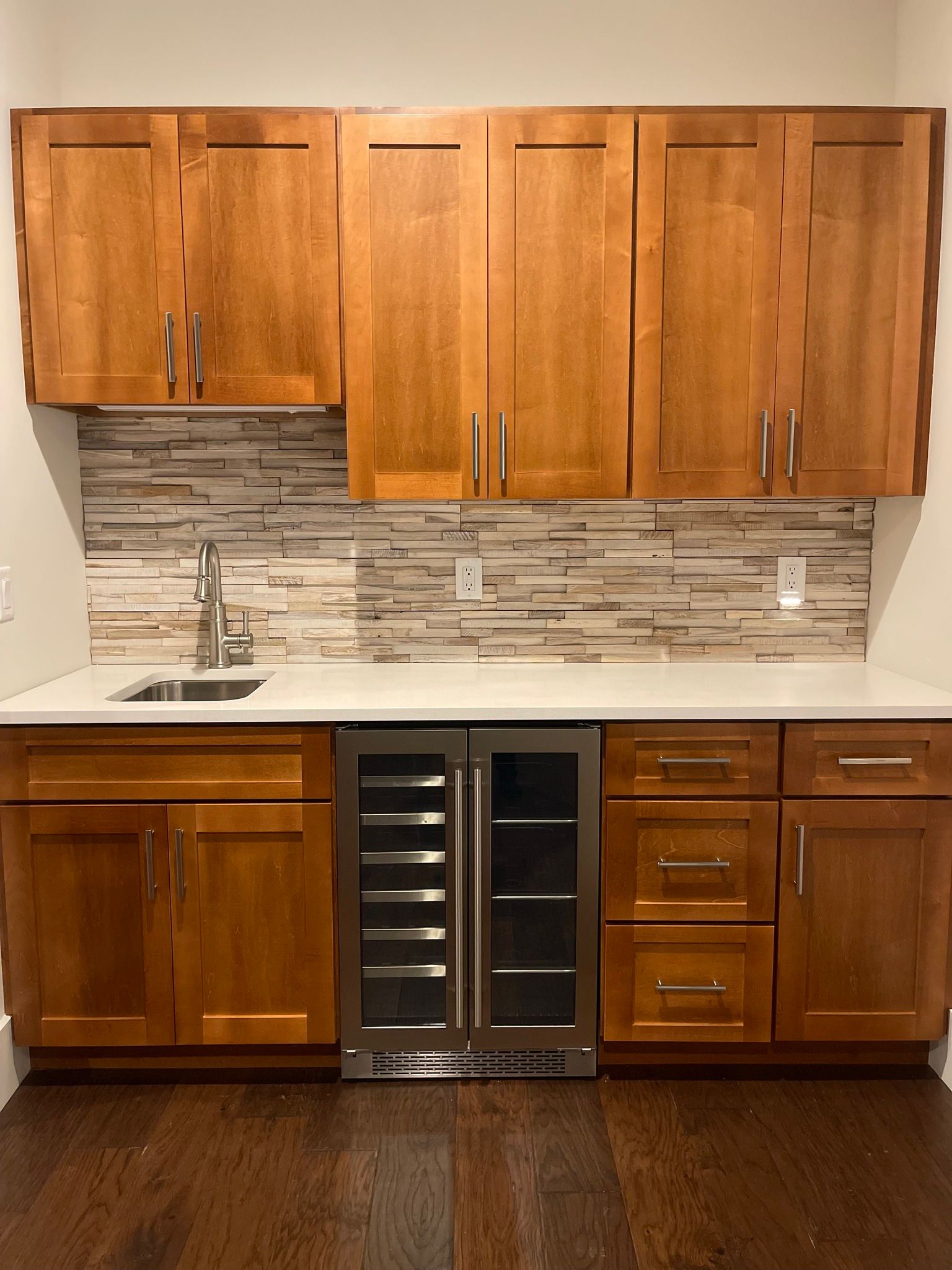 A wet bar featuring honey-toned wood cabinets, a white countertop, a stainless steel wine cooler, and a tiled backsplash.