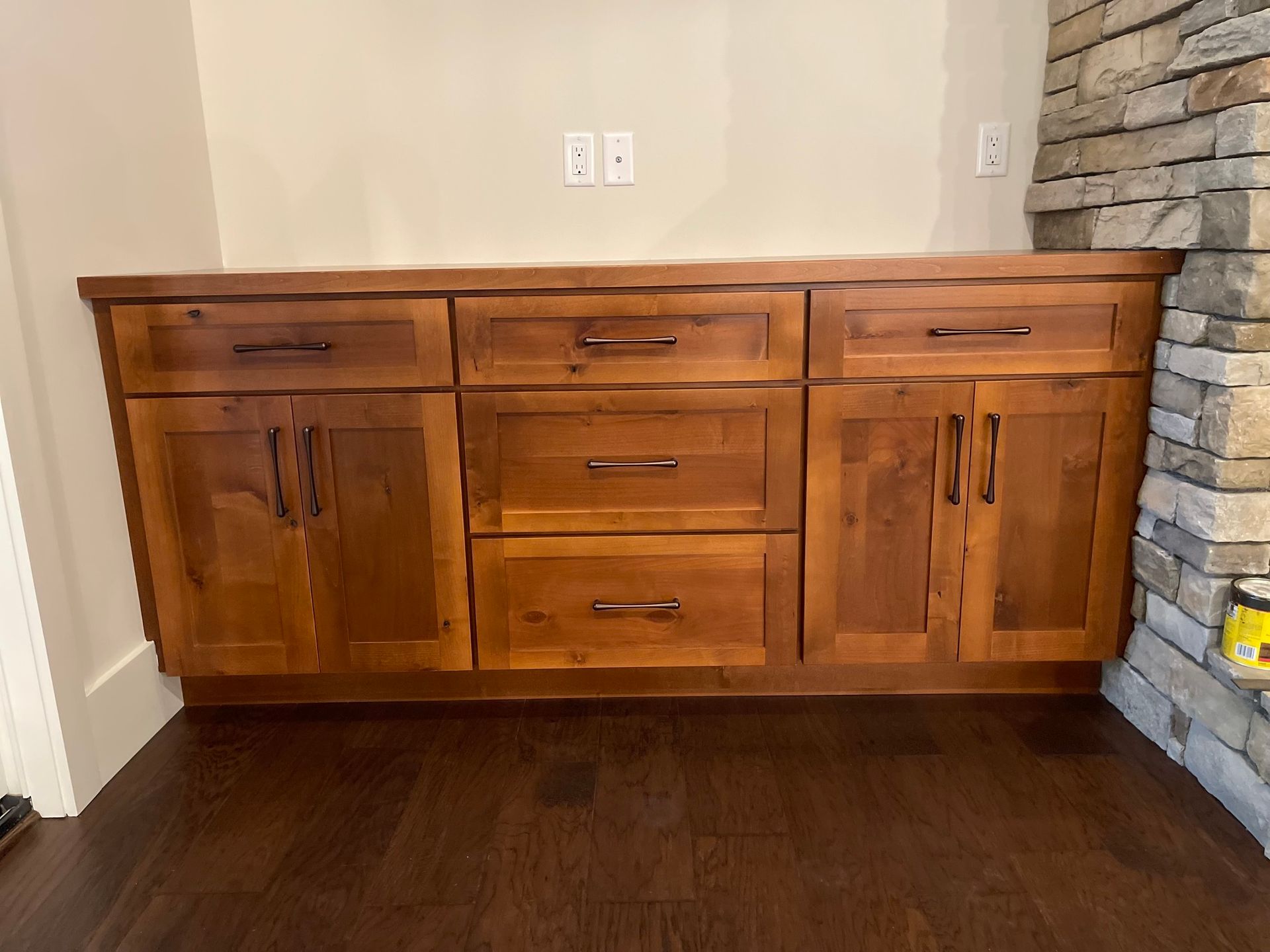 A wooden media cabinet with two side doors and three center drawers, set against a wall next to a stone fireplace.