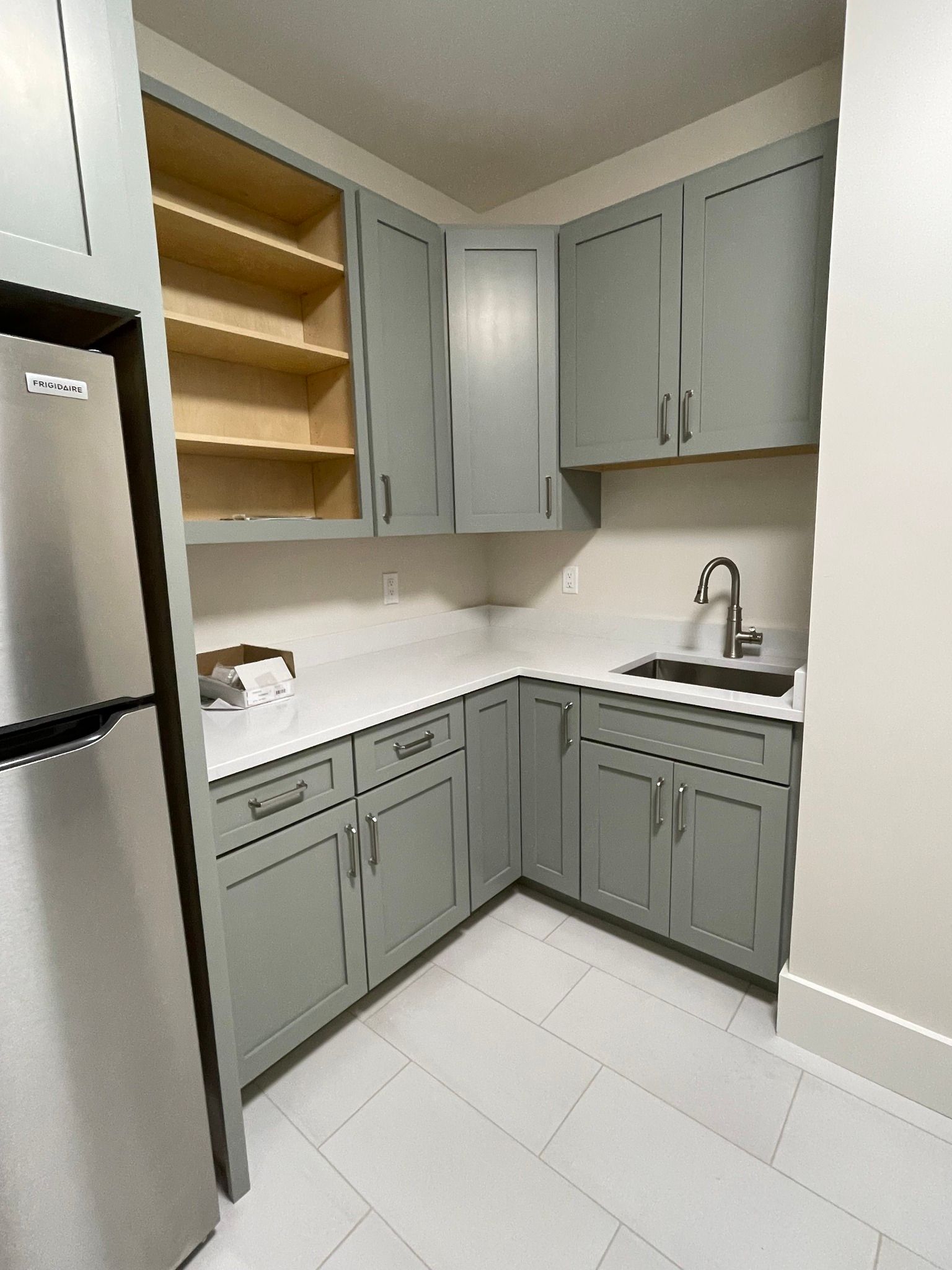 A small kitchen corner featuring sage green cabinetry, a stainless steel sink, white countertops, and light tiled floors.