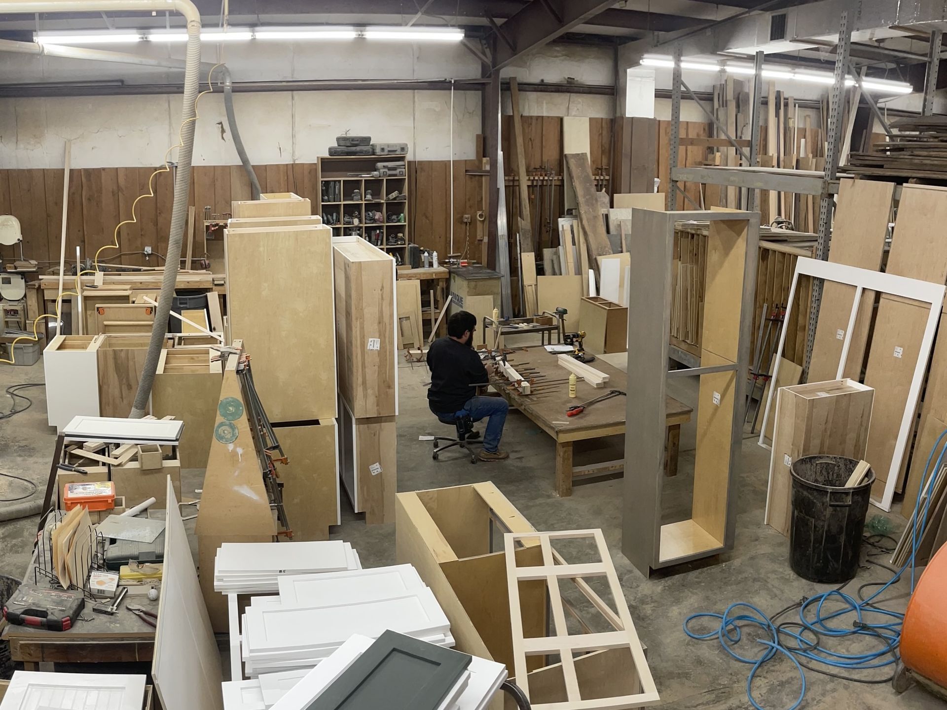 A woodworker sits at a workbench in a cluttered cabinet shop filled with wooden panels, frames, and tools.