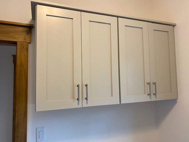Three light-colored Shaker-style wall cabinets with silver handles mounted on a white wall next to a wooden door frame.