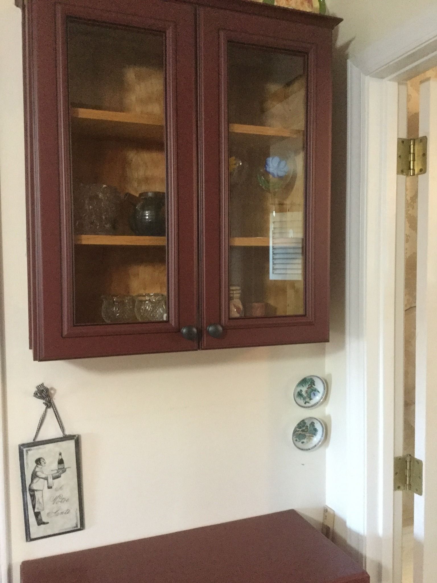 A maroon glass-front wall cabinet hangs above a counter, with a framed sign and two small decorative plates on the wall.
