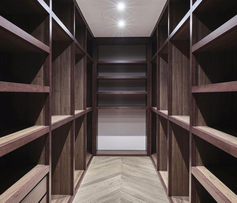 Walk-in closet with dark wood shelving units lining both sides and the back wall, featuring light herringbone wood floors.