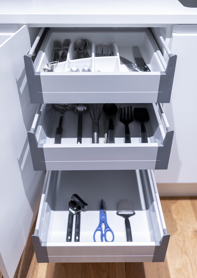 Three white kitchen drawers are fully extended, revealing organized cutlery, cooking utensils, and various tools.
