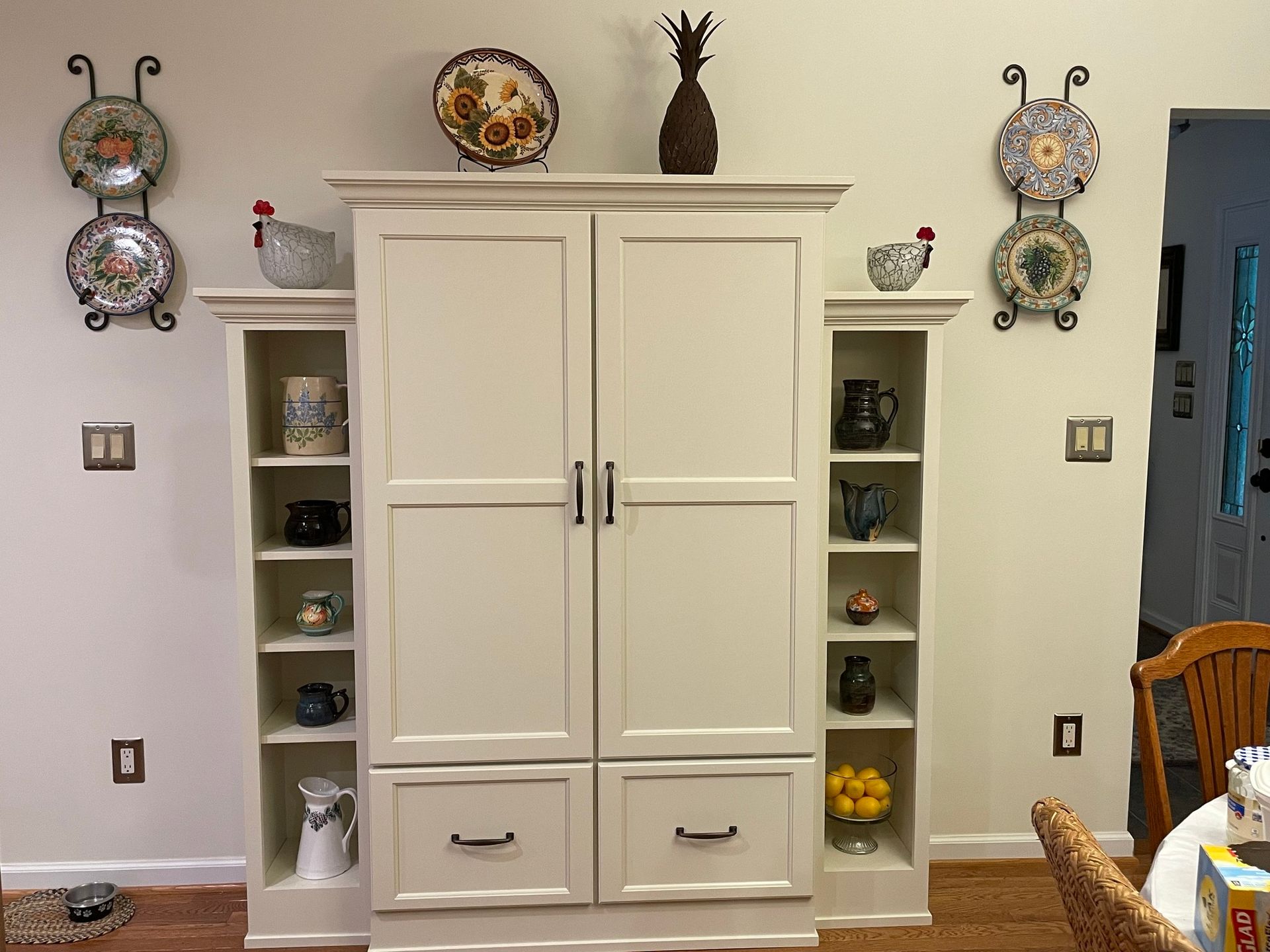 A tall, off-white cabinet with open shelves on either side, decorated with ceramic plates, figurines, and kitchen items.