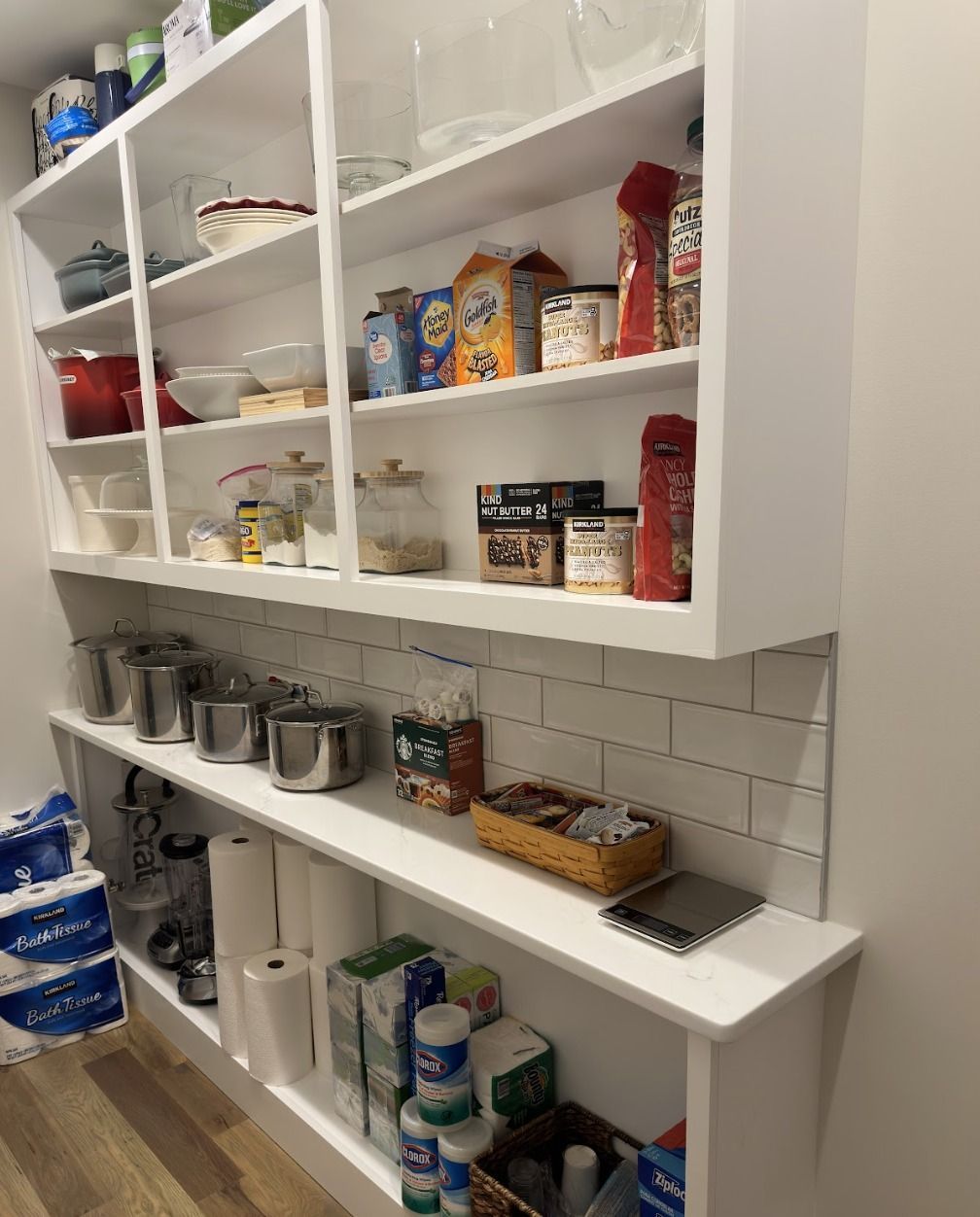 White pantry shelves with various food items, containers, cookware, and paper towels organized against a tiled wall.