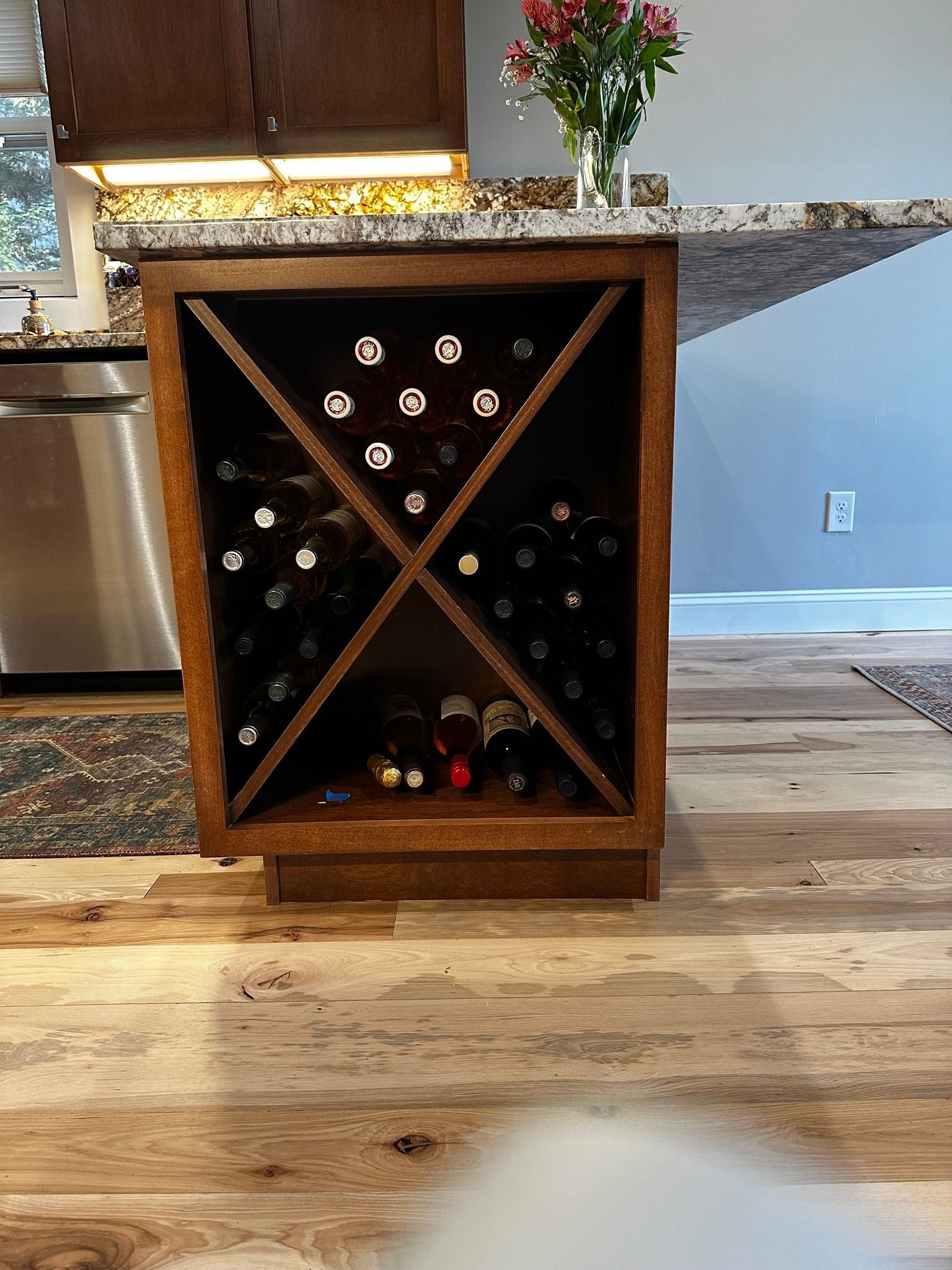 A wooden kitchen island end cabinet features X-shaped dividers storing multiple wine bottles on a hardwood floor.