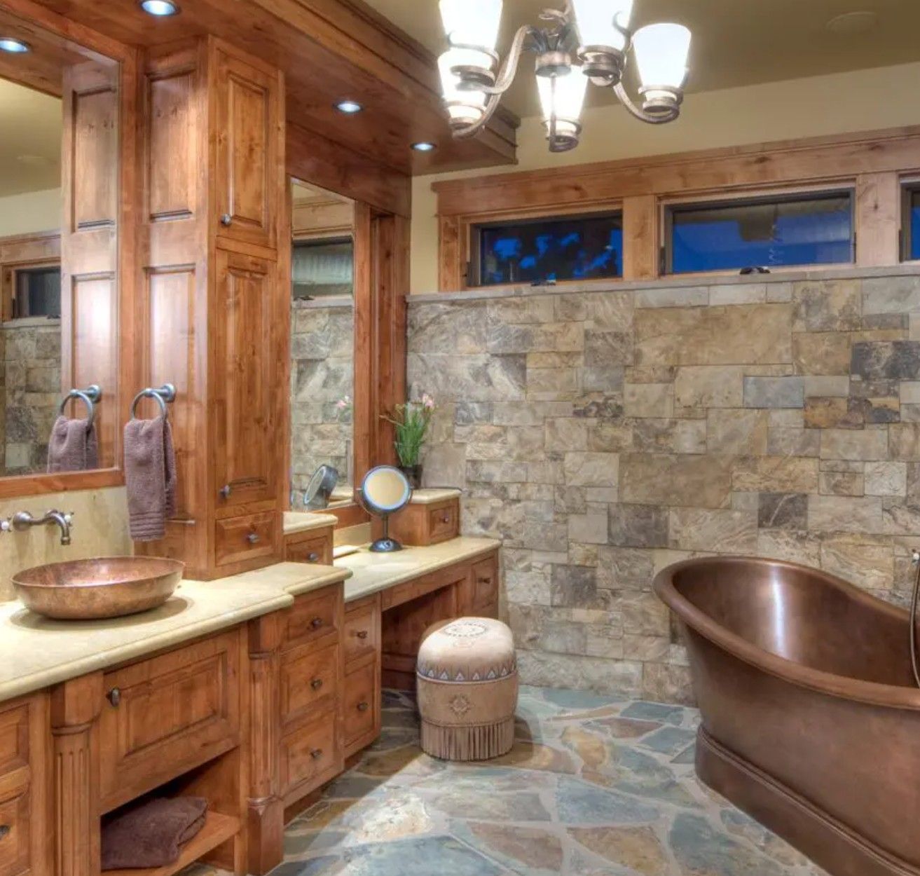 A rustic bathroom featuring stone walls, a copper soaking tub, a stone floor, and a wooden vanity with a copper basin sink.