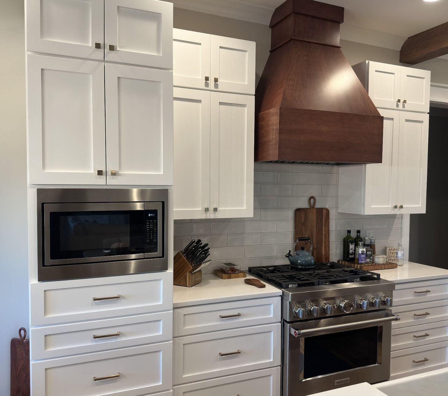 A modern kitchen with white Shaker cabinets, a large dark wood range hood, stainless steel appliances, and tile backsplash.