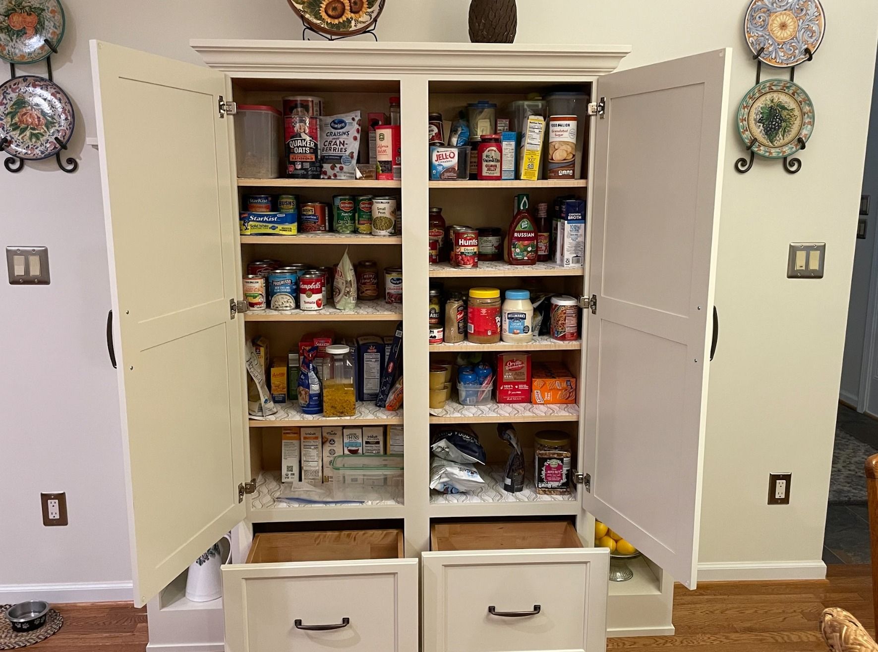 An open, cream-colored pantry cabinet filled with food items on shelves, featuring two lower storage drawers.