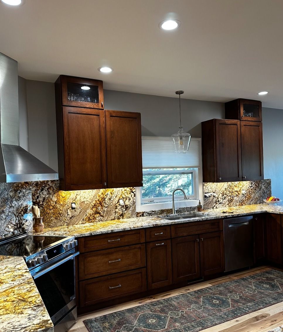 A kitchen with dark wood cabinets, granite countertops, an under-cabinet light, a stainless steel range, and a rug.