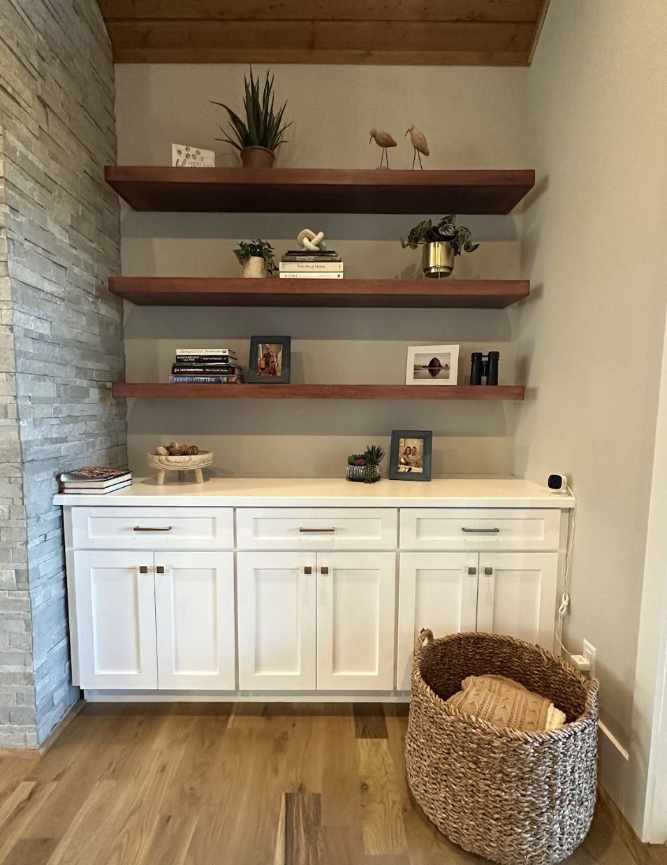 White cabinets topped with three wooden floating shelves featuring books, plants, and decor against a stone wall accent.