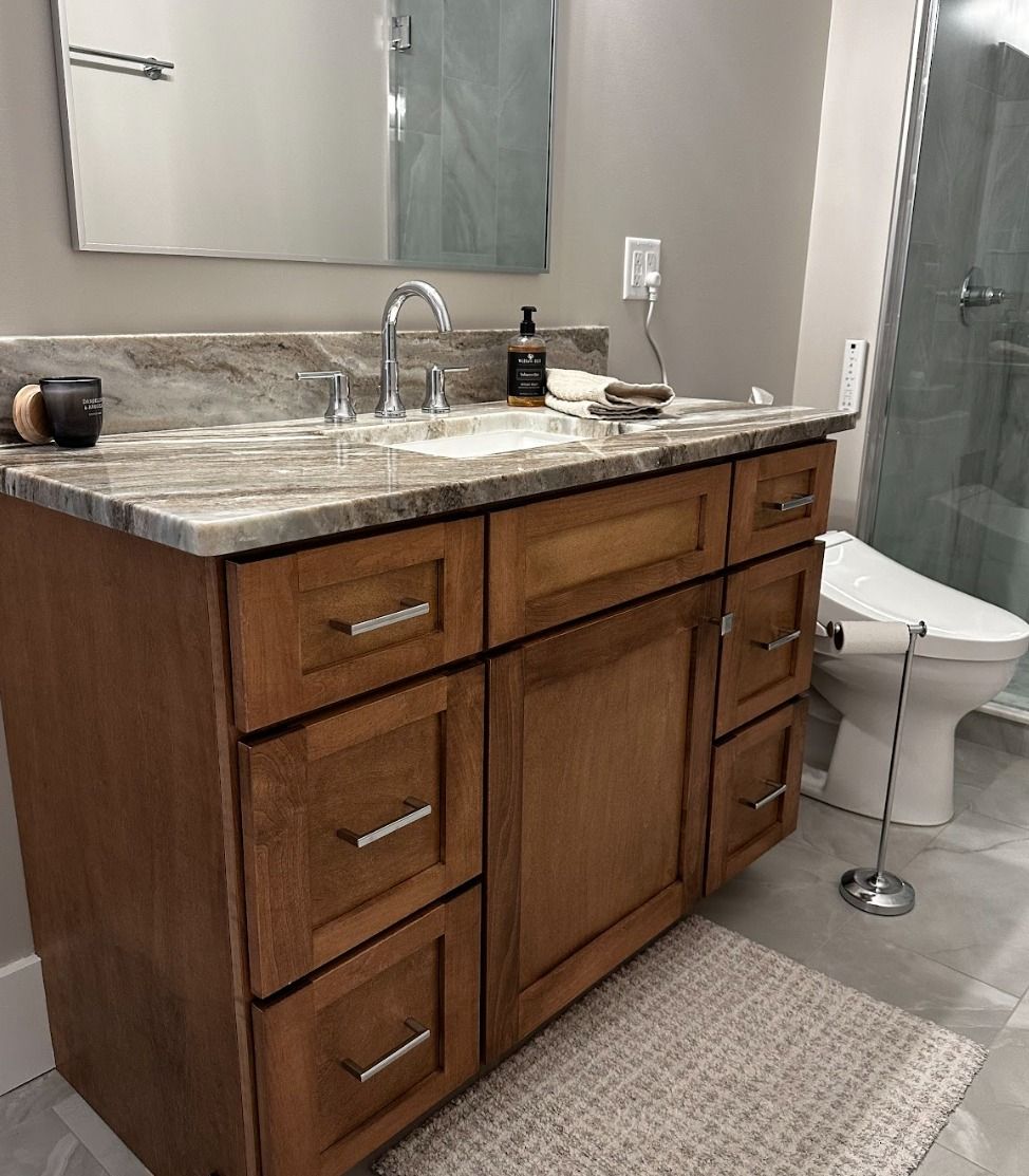 A wooden bathroom vanity with a granite countertop, sink, and mirror, positioned next to a toilet in a tiled bathroom.