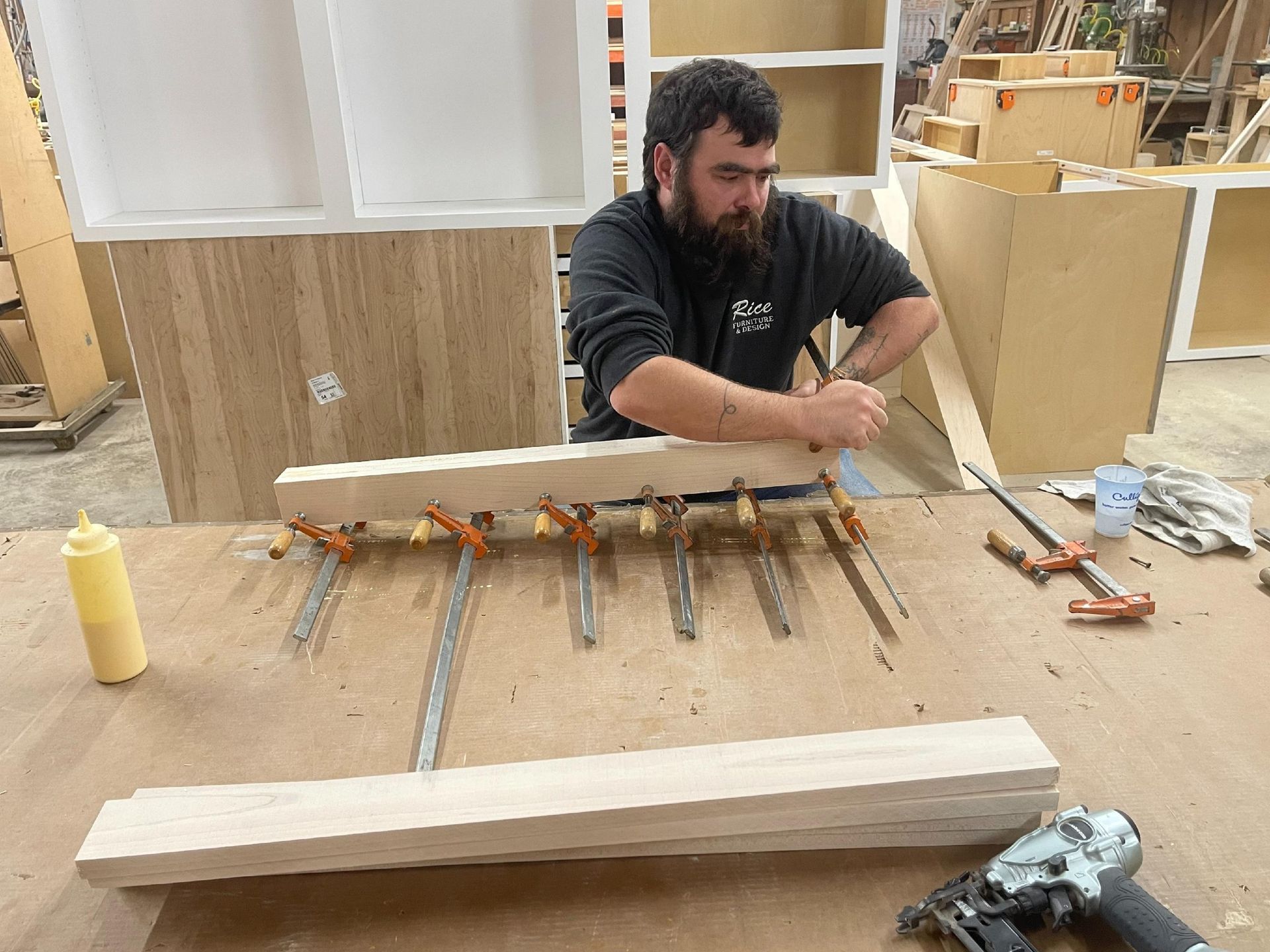 A person with a beard uses clamps to glue pieces of light wood together in a woodworking shop.