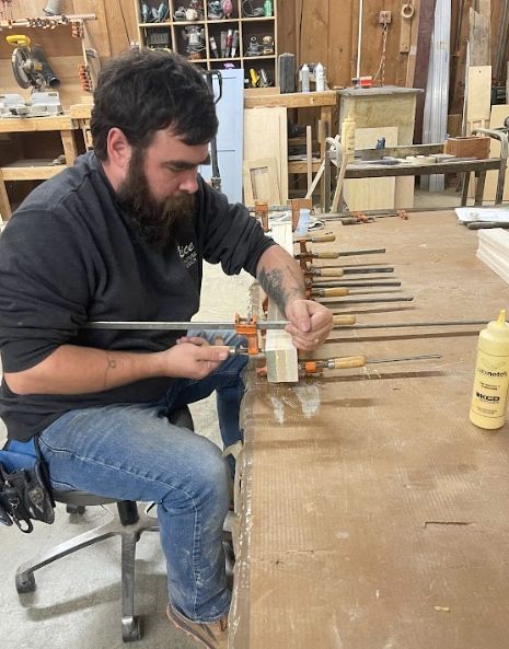 A person works at a woodworking shop, using clamps to glue wooden boards together on a large workbench.